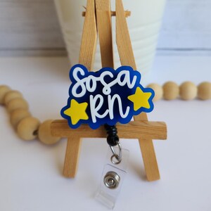 May include: A blue and yellow badge reel with the text "Sosa RN" in white script. The badge reel has two yellow star accents and a clear plastic retractable clip. The badge is displayed on a small wooden easel.