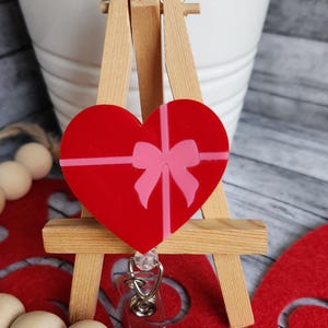 May include: Red heart-shaped badge holder with a pink ribbon and bow. The holder is attached to a retractable badge reel.