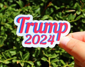 May include: A white sticker with the text "Trump 2024" in red and blue lettering. The sticker has a glossy finish and is slightly curved.