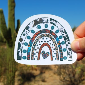 May include: A colorful rainbow sticker with a heart in the center. The rainbow is made up of different patterns, including cowhide, leopard print, and geometric designs. The sticker is on a white background.
