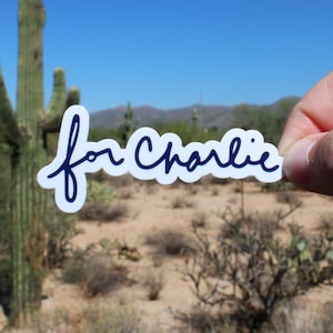 May include: A white sticker with the words "for Charlie" in a dark blue, handwritten font. The sticker is held up against a desert landscape with cacti and a clear blue sky.