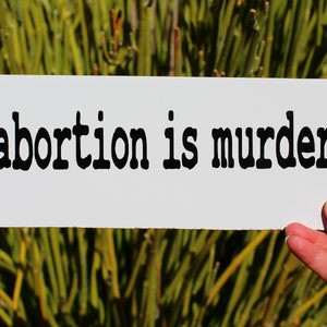 May include: A white sign with black text that reads "abortion is murder".