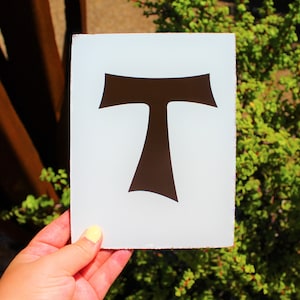 May include: A white rectangular sign featuring a dark brown stylized letter "T". The sign is held in front of a green leafy background and a rusty metal structure. The edges of the sign appear slightly distressed.