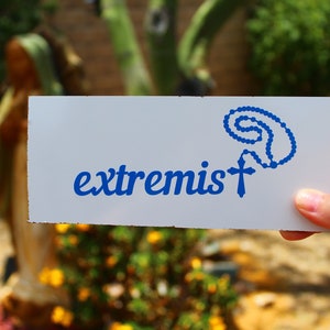 May include: A white rectangular sign with the word "extremist" in blue script, accompanied by a blue rosary and cross design. The sign is held up in front of a blurred outdoor background with greenery and yellow flowers.