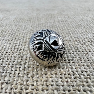 Könnte beinhalten: Ein silberner Metallknopf mit geometrischem Design. Der Knopf hat eine erhabene, sternförmige Mitte mit einem diamantartigen Muster.