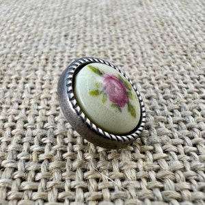 Op de afbeelding: Een zilverkleurige knoop met een witte porseleinen bovenkant met een roze roos en groene bladeren.