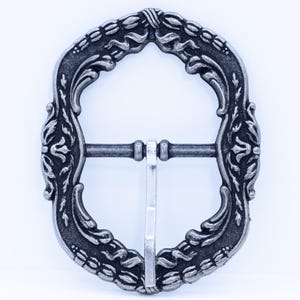 Könnte beinhalten: Eine silberfarbene Gürtelschnalle mit einem verzierten, floralen Design. Die Schnalle hat eine rechteckige Form mit abgerundeten Ober- und Unterseiten.