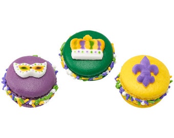 May include: Three colorful macarons with Mardi Gras-themed decorations. One is purple with a mask, another is green with a crown, and the last is yellow with a fleur-de-lis. Each is decorated with sprinkles.