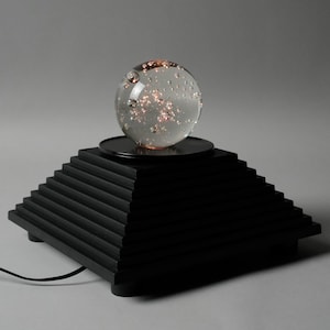 Op de afbeelding: Decoratieve lamp met een heldere glazen bol met interne verlichting, rustend op een zwarte, trapsgewijze piramidevormige basis. De lamp heeft een zwart netsnoer. Neutrale achtergrond.