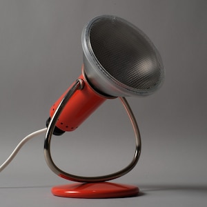 May include: A retro-style desk lamp with a vibrant orange body and a silver-colored, mesh-covered light. The lamp features a curved, metallic stand and a white power cord. The lamp's base is a matching orange color.