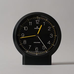 Könnte beinhalten: Eine schwarze, runde Tischuhr mit schwarzem Zifferblatt, weißen arabischen Ziffern sowie gelben Stunden- und Minutenzeigern. Die Uhr zeigt den Schriftzug JUNGHANS und radiocontrolled, ergänzt durch einen dünnen gelben Kreis und einen schwarzen Sockel.