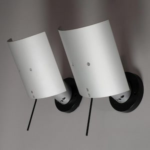 Könnte beinhalten: Zwei weiße, zylindrische Wandleuchten mit schwarzen, kreisförmigen Basen und schwarzen Stielen. Die Leuchten sind an einer grauen Wand montiert. Modernes, minimalistisches Design.