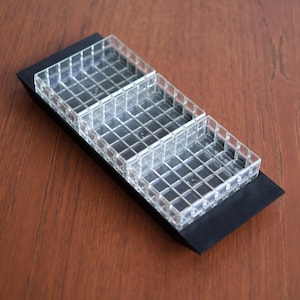 Op de afbeelding: Een heldere, rechthoekige organizer met drie compartimenten, geplaatst op een zwarte basis. De compartimenten zijn verdeeld door een rasterpatroon. De organizer staat op een bruin houten oppervlak.