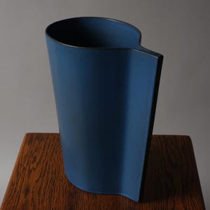 Puede incluir: Un jarrón azul moderno con un diseño asimétrico único. El jarrón tiene un borde oscuro y una forma curva, colocado sobre una superficie de madera. La altura del jarrón es de aproximadamente 30 cm.