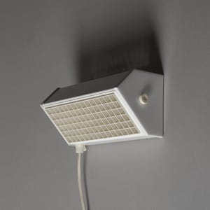 May include: A white, geometric wall-mounted light fixture with a grid-like front panel. The light has a dark gray top and a white power cord. It is mounted on a gray wall.