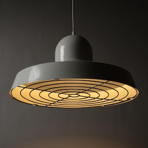Peut inclure: Suspension luminaire grise avec un motif de grille circulaire en dessous. Le luminaire a un dessus arrondi et un abat-jour large et plat. Il est suspendu à un seul cordon. Idéal pour une cuisine ou une salle à manger.