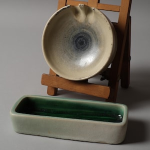 Może przedstawiać: Ceramiczna popielniczka z jasnobeżowym i niebieskim wzorem wirowym, spoczywająca na małym drewnianym sztaludze. Poniżej prostokątna, jasnozielona ceramiczna miska z ciemnozielonym wnętrzem.