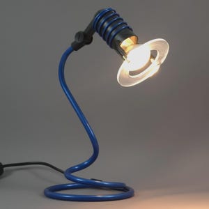May include: A blue and black desk lamp with a unique, curved design. The lamp features a circular base, a flexible neck, and a spotlight with a clear, circular shade. The lamp is turned on, emitting a warm, inviting glow.
