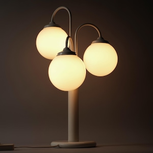 Op de afbeelding: Een tafellamp met een centrale verticale paal, een ronde voet en drie gebogen armen die naar boven reiken. Elke arm draagt een bolvormige, matwitte glazen kap die een warm, zacht licht uitstraalt. Een kleine rechthoekige schakelaar is bevestigd.