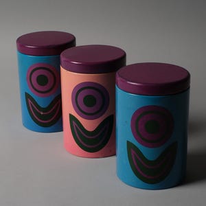 May include: Three cylindrical containers with lids. The containers are painted in blue, pink, and blue. Each container has a purple lid and a decorative design of concentric circles and a curved shape in black and green.
