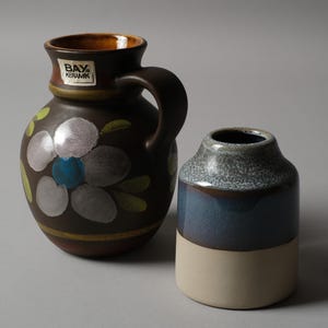 May include: Two ceramic vases. One is a brown pitcher with a handle, floral design, and the text "BAY KERAMIK". The other is a small, cylindrical vase with a blue and cream color scheme.