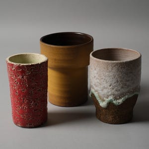 Può includere: Tre vasi in ceramica di varie dimensioni e trame. Il più alto è un vaso cilindrico giallo senape. Quello centrale è un cilindro rosso strutturato. Il terzo è un cilindro bianco e marrone strutturato.