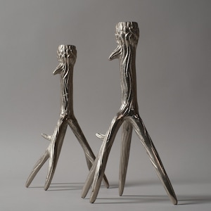 May include: Two silver-coloured, tree-branch-shaped candle holders. The candle holders have a textured surface and a three-legged base. The top of the candle holders are designed to hold a single candle. The background is a solid grey.