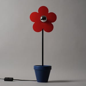 Puede incluir: Una lámpara con forma de flor que presenta una flor roja brillante de cinco pétalos con una esfera central cromada, sostenida por un tallo negro fino que surge de una maceta azul sólida. Un cable eléctrico negro con interruptor sale de la base.