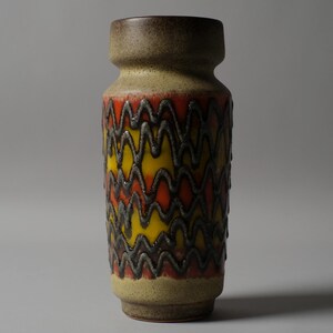 May include: A ceramic vase with a textured, zig-zag pattern in yellow, orange, and brown. The vase has a cylindrical shape with a slightly flared top and base. The height is approximately 10 inches.