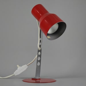 May include: A red desk lamp with a silver metal arm and base. The lamp has a conical shade and a white cord with a switch. The lamp is on a gray background.