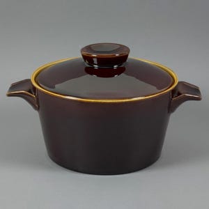 May include: A dark brown ceramic casserole dish with a matching lid. The lid has a small, round handle and a gold-colored rim. The dish has two handles on either side.
