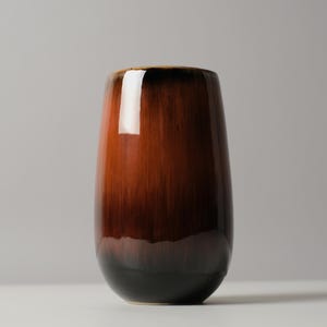 May include: A cylindrical ceramic vase with a smooth, glossy finish. The color transitions from a deep, dark brown at the base to a lighter, warm reddish-brown toward the top, featuring a reflective surface with a prominent light highlight.