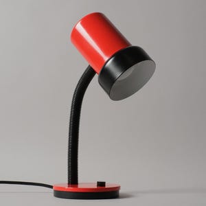Könnte beinhalten: Eine Schreibtischlampe mit einem zylindrischen roten Kopf, einem schwarzen Rand und einem weißen Inneren. Die Lampe hat einen flexiblen schwarzen Schwanenhalsarm, der an einem runden roten Sockel mit schwarzem Boden und kleinem schwarzen Schalter befestigt ist.