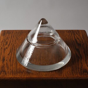 The Crystalline Silhouette. This second image, captured from a high angle on an oak surface, demonstrates the object's remarkable optical clarity. The solid crystal apex acts as a prism, gathering ambient light and refracting it through the thick walls of the vessel. The absence of any surface decoration allows the pure geometric relationship between the conical lid and the flared base to remain the focal point.