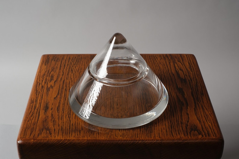 The Crystalline Silhouette. This second image, captured from a high angle on an oak surface, demonstrates the object's remarkable optical clarity. The solid crystal apex acts as a prism, gathering ambient light and refracting it through the thick walls of the vessel. The absence of any surface decoration allows the pure geometric relationship between the conical lid and the flared base to remain the focal point.