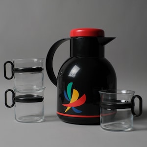 Puede incluir: Una jarra térmica negra con tapa roja, asa negra y diseño abstracto colorido. Tres tazas de vidrio transparente con asas y bandas negras apiladas cerca, con otra taza a la derecha.