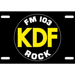 May include: Black license plate with a white circle containing the text "FM 103 KDF ROCK" in white and yellow lettering.