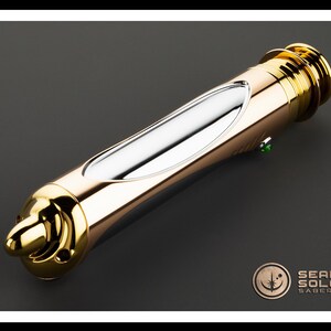 May include: A gold and silver lightsaber with a green button. The lightsaber has a sleek design with a metallic finish. The product is from Sean Solo Sabers.