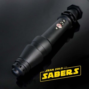 Może przedstawiać: Czarna rękojeść miecza świetlnego z srebrnym przyciskiem i czerwoną diodą LED. Rękojeść jest wykonana z metalu i ma fakturowaną powierzchnię. Rękojeść jest oznaczona napisem "Sean Solo Store Sabers".