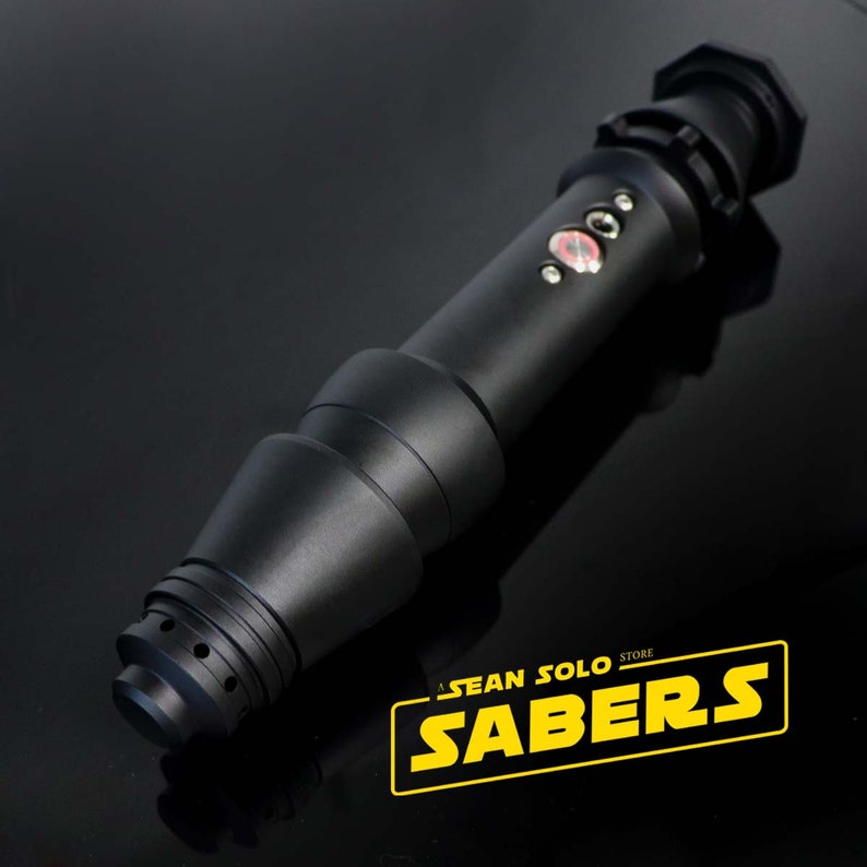 Może przedstawiać: Czarna rękojeść miecza świetlnego z srebrnym przyciskiem i czerwoną diodą LED. Rękojeść jest wykonana z metalu i ma fakturowaną powierzchnię. Rękojeść jest oznaczona napisem "Sean Solo Store Sabers".