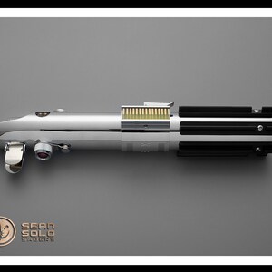 Puede incluir: Un accesorio de sable de luz cromado con detalles en negro y plata. El accesorio tiene un dise&ntilde;o cil&iacute;ndrico con una serie de crestas negras y un mango plateado. El texto "SEAN SOLO SABERS" es visible en la parte inferior izquierda.