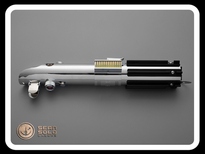 Puede incluir: Un accesorio de sable de luz cromado con detalles en negro y plata. El accesorio tiene un dise&ntilde;o cil&iacute;ndrico con una serie de crestas negras y un mango plateado. El texto "SEAN SOLO SABERS" es visible en la parte inferior izquierda.