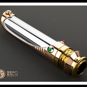 May include: A metallic lightsaber with gold and silver accents, featuring a green and red button. The hilt has a polished finish, and the design includes the text "SEAN SOLO SABERS" and a logo.