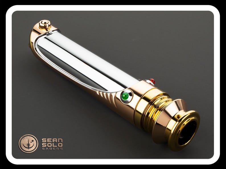 May include: A metallic lightsaber with gold and silver accents, featuring a green and red button. The hilt has a polished finish, and the design includes the text "SEAN SOLO SABERS" and a logo.