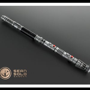 May include: A metallic silver lightsaber hilt with a long, cylindrical body featuring intricate perforated patterns, ribbed sections, and two small red circular buttons. The hilt includes a logo with the text SEAN SOLO SABERS in the corner.