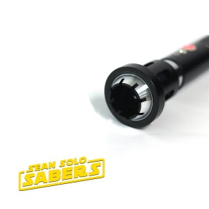 May include: Close-up of the end of a black lightsaber hilt with a silver ring around the opening. The hilt has a textured surface and a small red dot on the side.