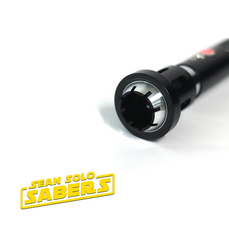 May include: Close-up of the end of a black lightsaber hilt with a silver ring around the opening. The hilt has a textured surface and a small red dot on the side.