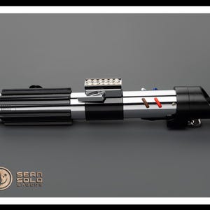 Op de afbeelding: Een zwart en zilveren lightsaber met een rode en bruine accent. De lightsaber heeft een reeks kleine lampjes aan de bovenkant.