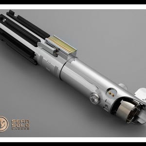 Puede incluir: Un detallado sable de luz plateado con detalles en negro y dorado. El accesorio presenta un dise&ntilde;o complejo con varios botones, interruptores y un emisor transparente. El logotipo de Sean Solo Sabers es visible en la esquina inferior izquierda.