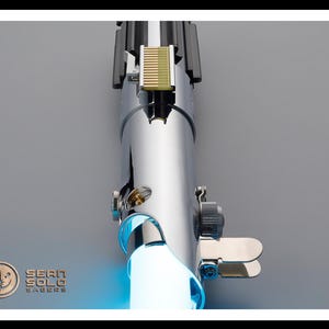 Puede incluir: Un sable de luz cromado con una hoja azul brillante. El mango presenta detalles intrincados, incluyendo una secci&oacute;n dorada y varios componentes met&aacute;licos. El logotipo "Sean Solo Sabers" es visible en la esquina inferior izquierda.